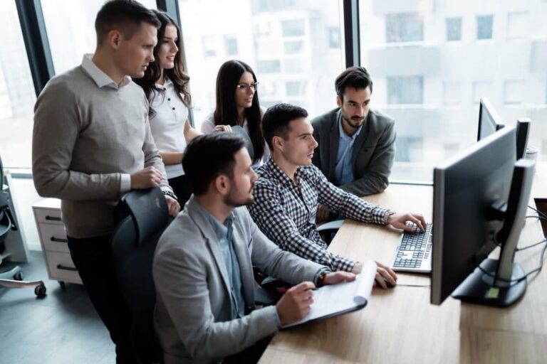 Eine Gruppe junger Berufstätiger versammelte sich in einer Büroumgebung um einen Schreibtisch und betrachtete gemeinsam einen Computermonitor.