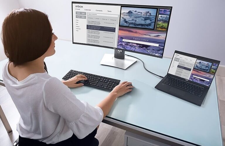 Person an einem Arbeitsplatz mit zwei Bildschirmen: einem großen Monitor und einem angeschlossenen Laptop auf einem Glastisch, auf denen E-Mails und Fotos nebeneinander angezeigt werden.