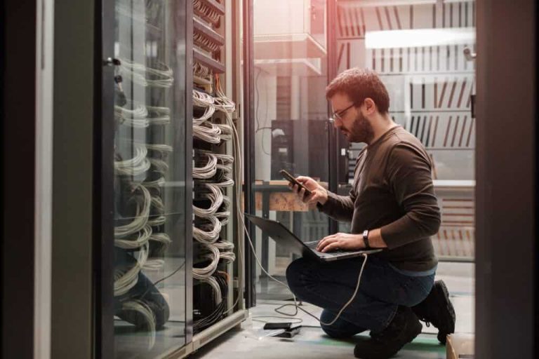 Technician kneeling beside server racks in a data centre, using a laptop and smartphone.