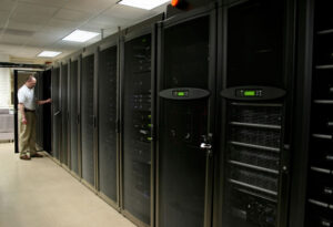 technician_working - A DataRoad - A DataRoad IT technician in a white shirt stands by a row of tall black server racks in a data center corridor, door open to a cabinet.