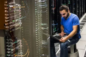 wireless - A DataRoad - A DataRoad Tech worker in a blue shirt sits beside server racks, using a laptop in a data center to troubleshoot.